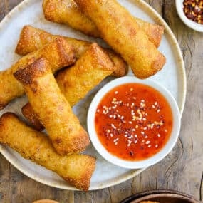 Sesame chicken egg rolls on a plate alongside a bowl of sweet chili dipping sauce. Small bowls of sliced scallions, sesame seeds and chili flakes surround the plate.