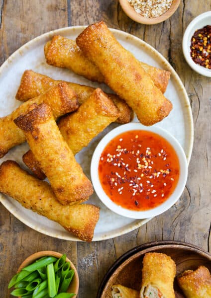 Sesame chicken egg rolls on a plate alongside a bowl of sweet chili dipping sauce. Small bowls of sliced scallions, sesame seeds and chili flakes surround the plate.