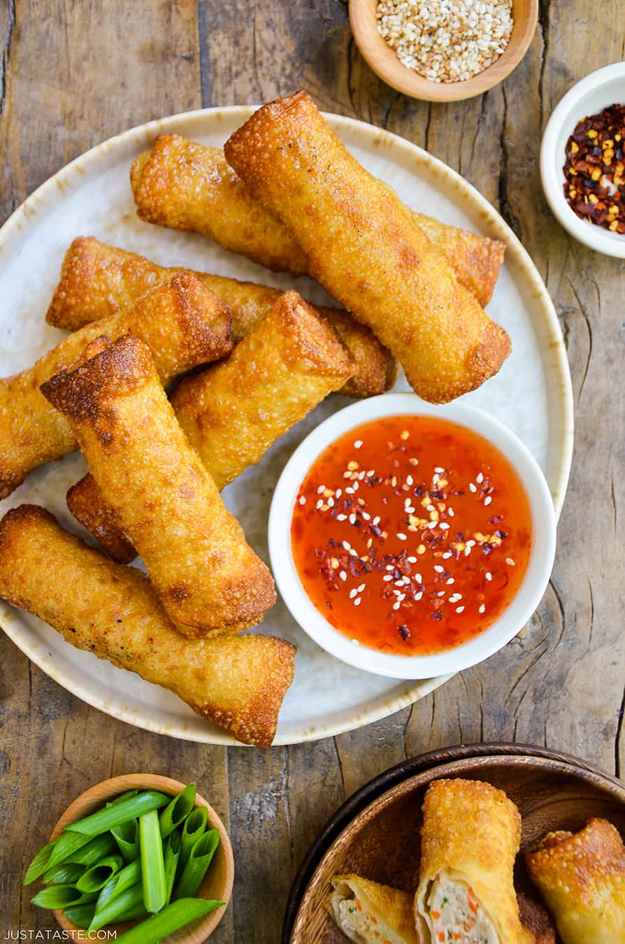 Sesame chicken egg rolls on a plate alongside a bowl of sweet chili dipping sauce. Small bowls of sliced scallions, sesame seeds and chili flakes surround the plate.