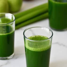 Glasses of green juice sit on a white marble countertop with celery and apples in the background.
