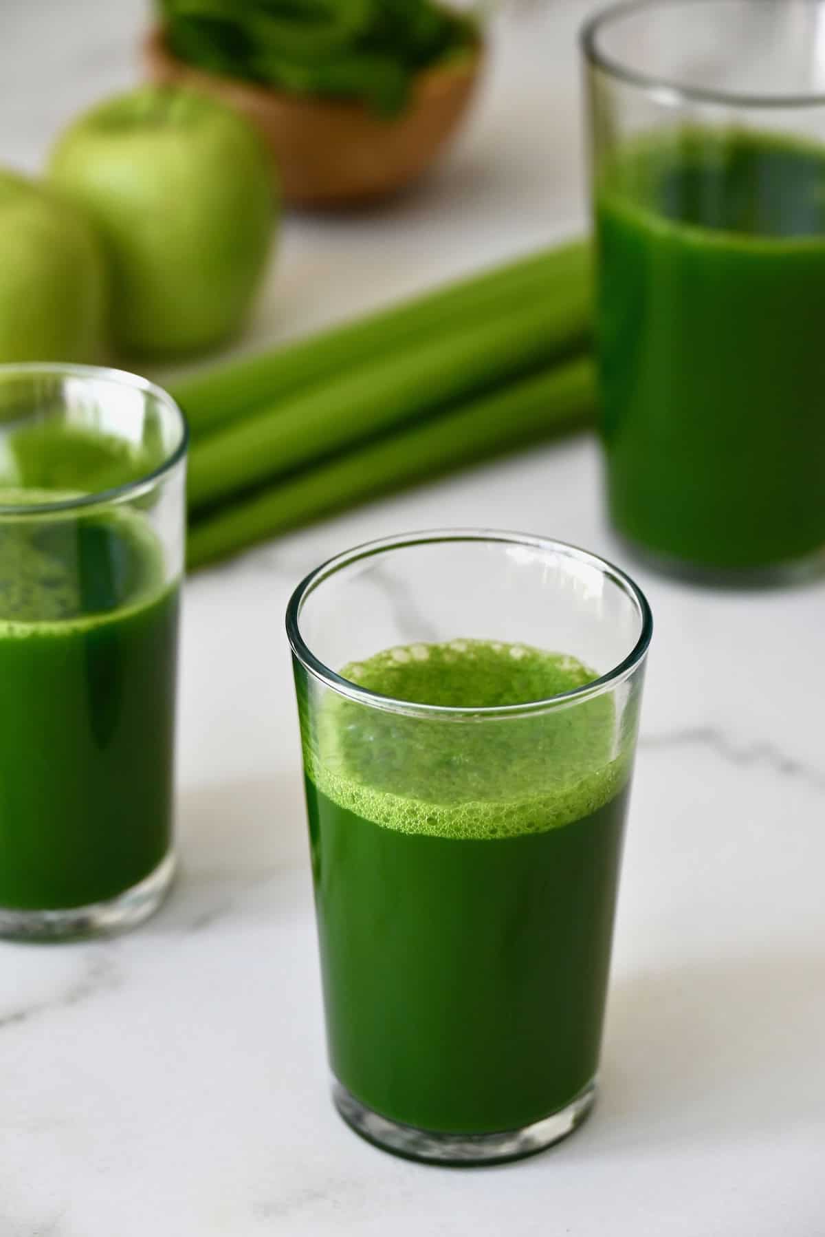 Glasses of green juice sit on a white marble countertop with celery and apples in the background.