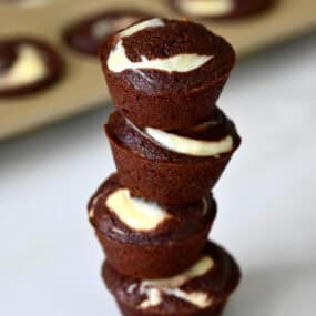 Cheesecake brownie bites are stacked on a marble counter, with a mini muffin pan in the background.