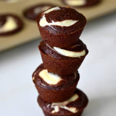 Cheesecake brownie bites are stacked on a marble counter, with a mini muffin pan in the background.