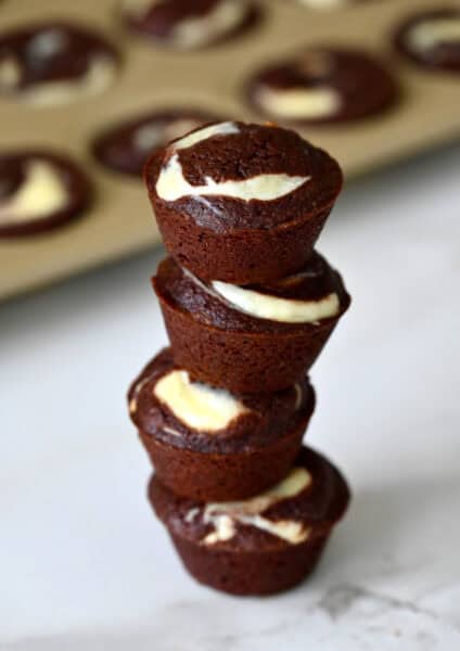 Cheesecake brownie bites are stacked on a marble counter, with a mini muffin pan in the background.