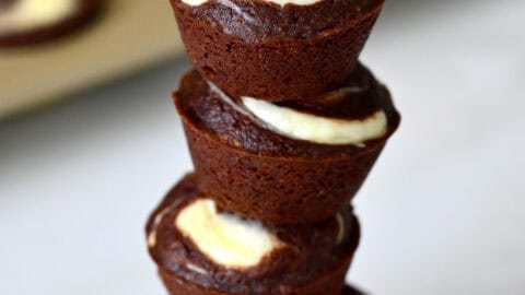 Cheesecake brownie bites are stacked on a marble counter, with a mini muffin pan in the background.