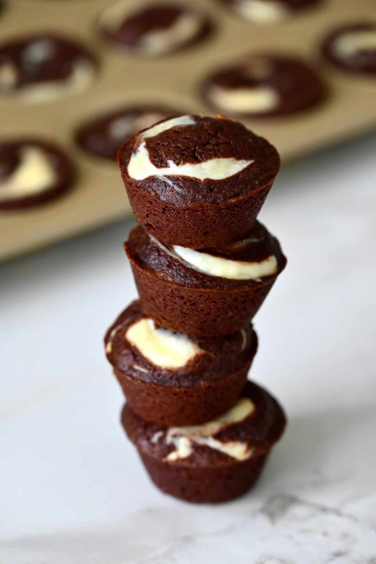 Cheesecake brownie bites are stacked on a marble counter, with a mini muffin pan in the background.