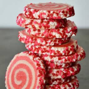 Pink pinwheel sugar cookies, edged in sprinkles, are stacked on a dark surface.