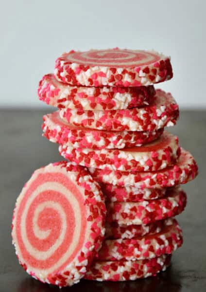Pink pinwheel sugar cookies, edged in sprinkles, are stacked on a dark surface.