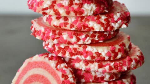 Pink pinwheel sugar cookies, edged in sprinkles, are stacked on a dark surface.
