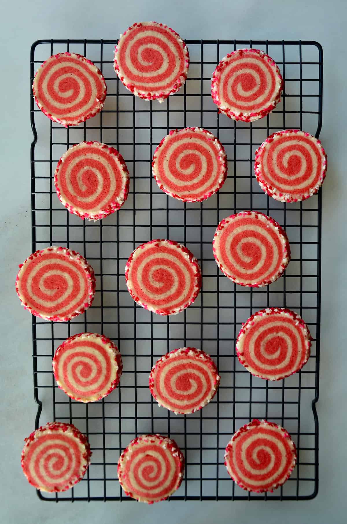 Pink pinwheel sugar cookies, edged in sprinkles, are arranged on a baking rack.