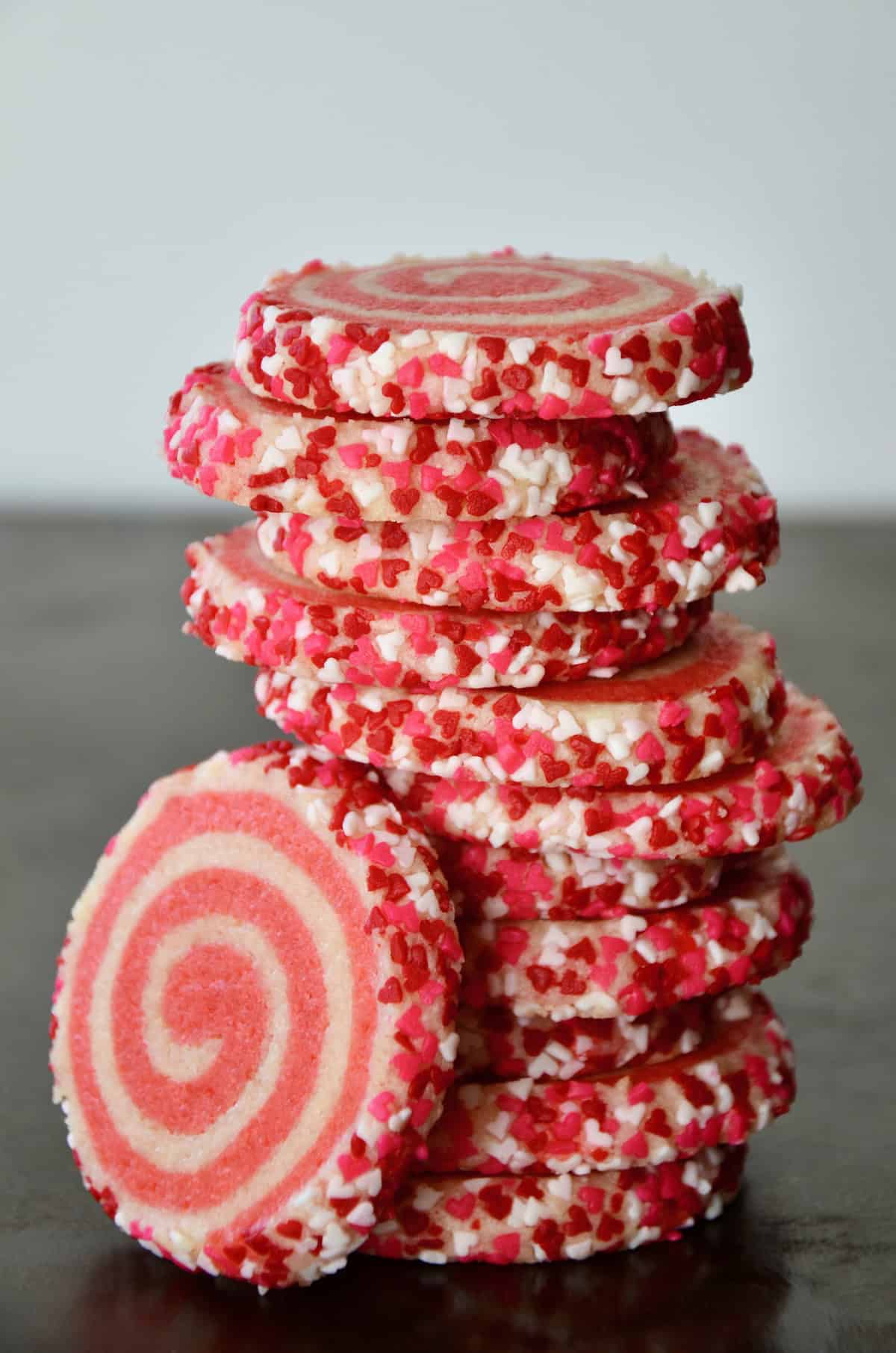 Pink pinwheel sugar cookies, edged in sprinkles, are stacked on a dark surface.