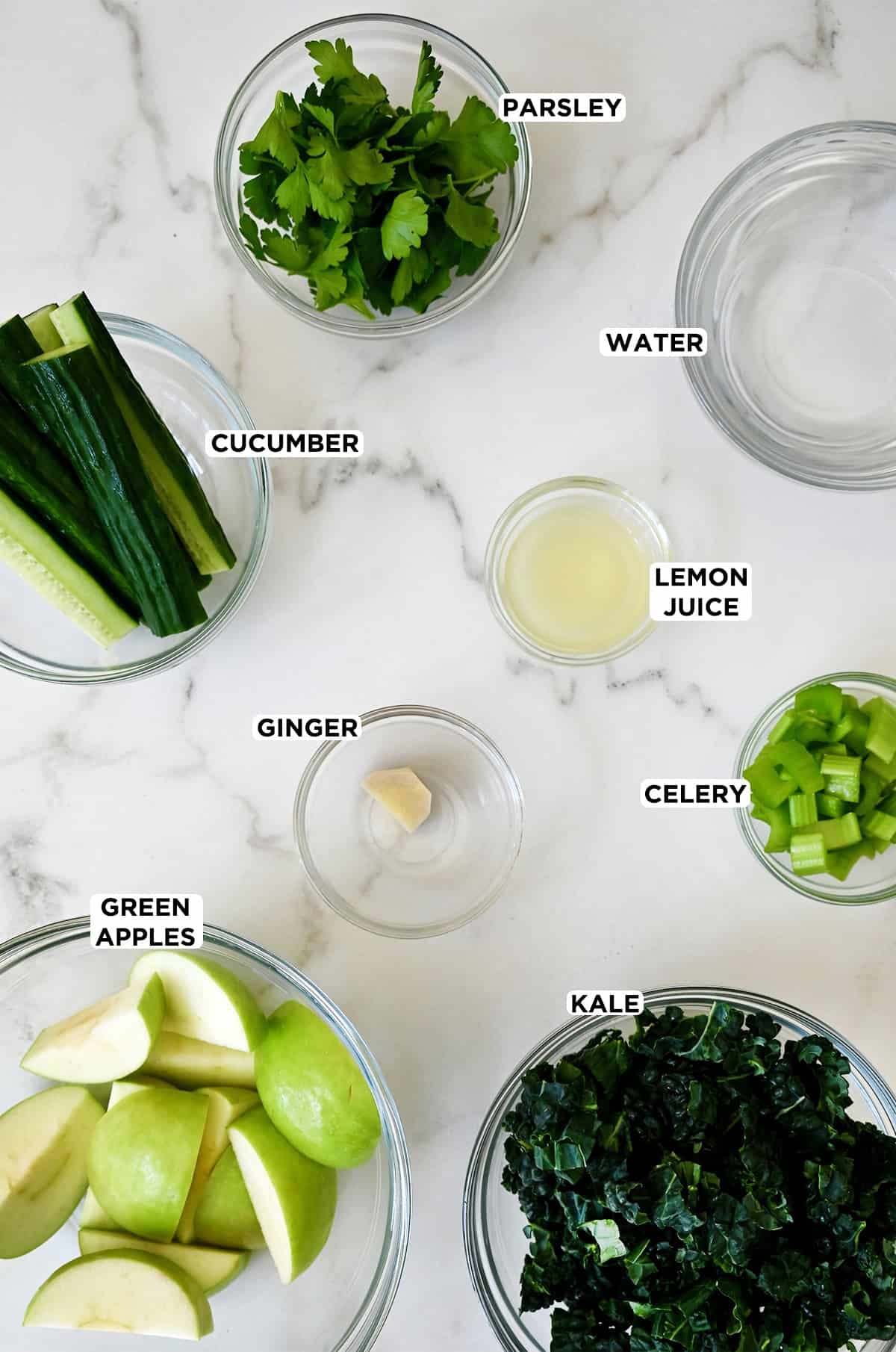 Glass bowls are filled with all the ingredients for green juice, including apples, kale, ginger, celery, parsley, cucumber and water.