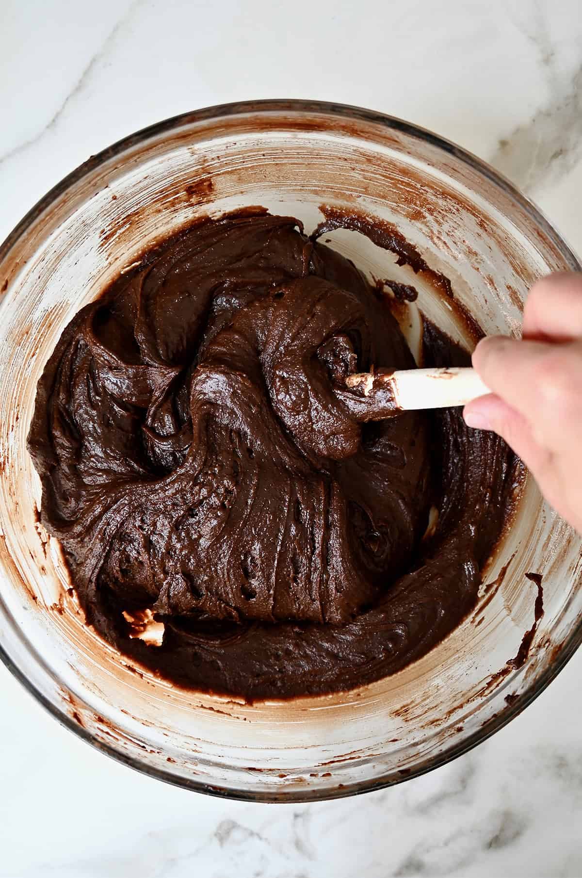 Brownie batter is being mixed with a spatula in a glass bowl.