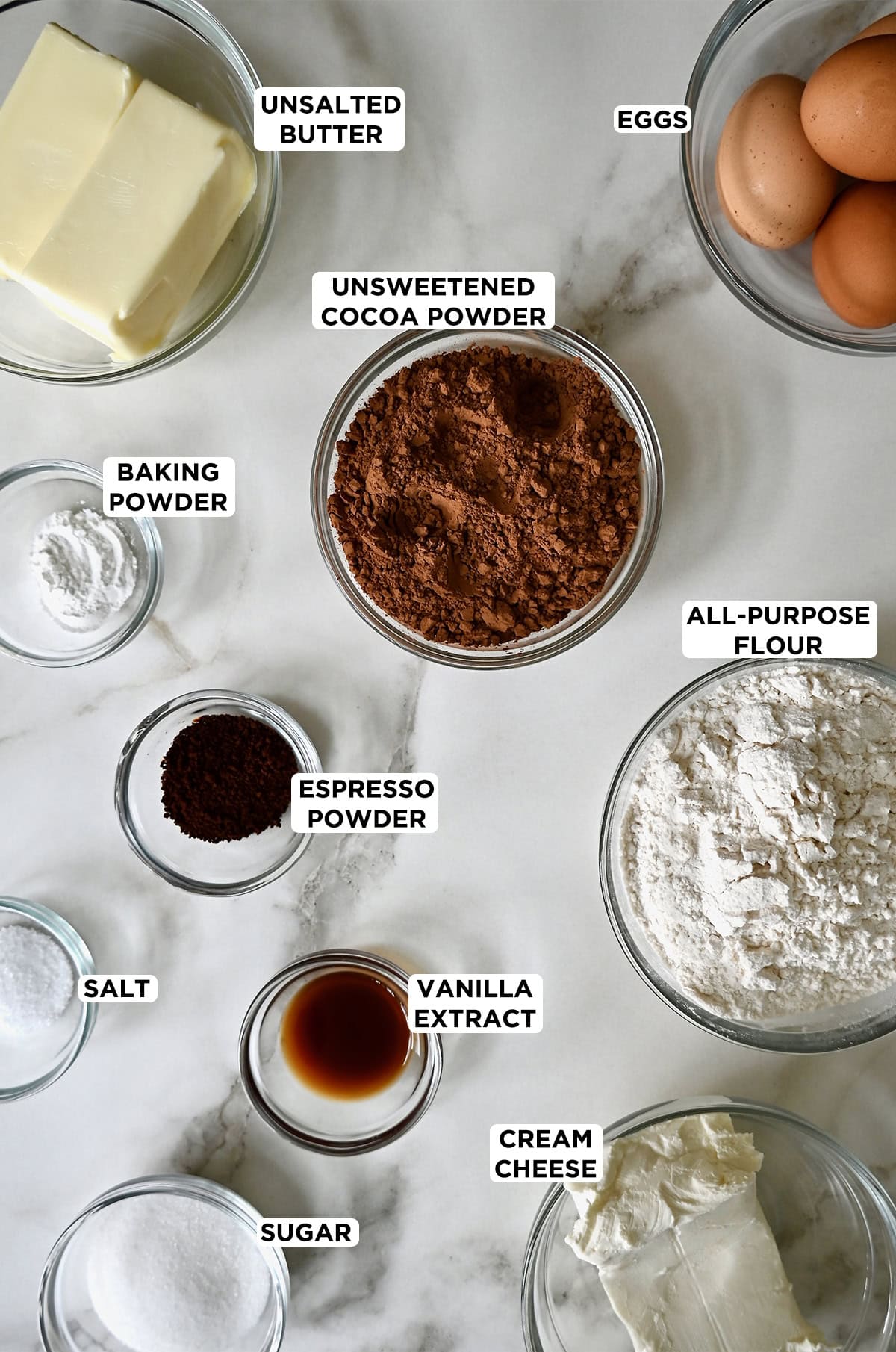 Clear glass bowls are filled with the ingredients for cheesecake brownie bikes, including sugar, cream cheese, vanilla, flour, salt, espresso powder, baking powder, butter, eggs and cocoa powder.