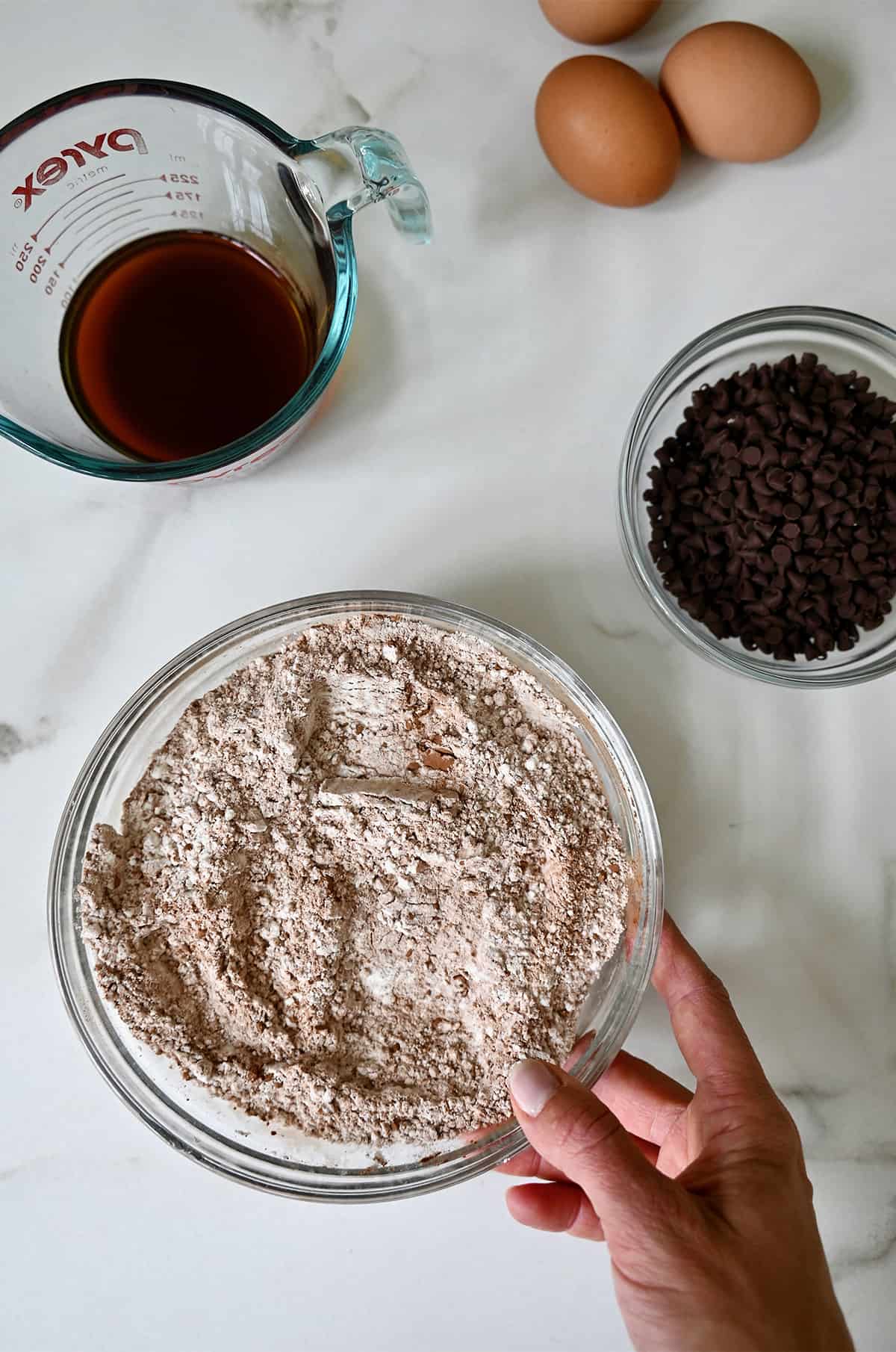 A medium bowl containing dry cake mix ingredients next to a small bowl with mini chocolate chips and a liquid measuring cup with coffee.