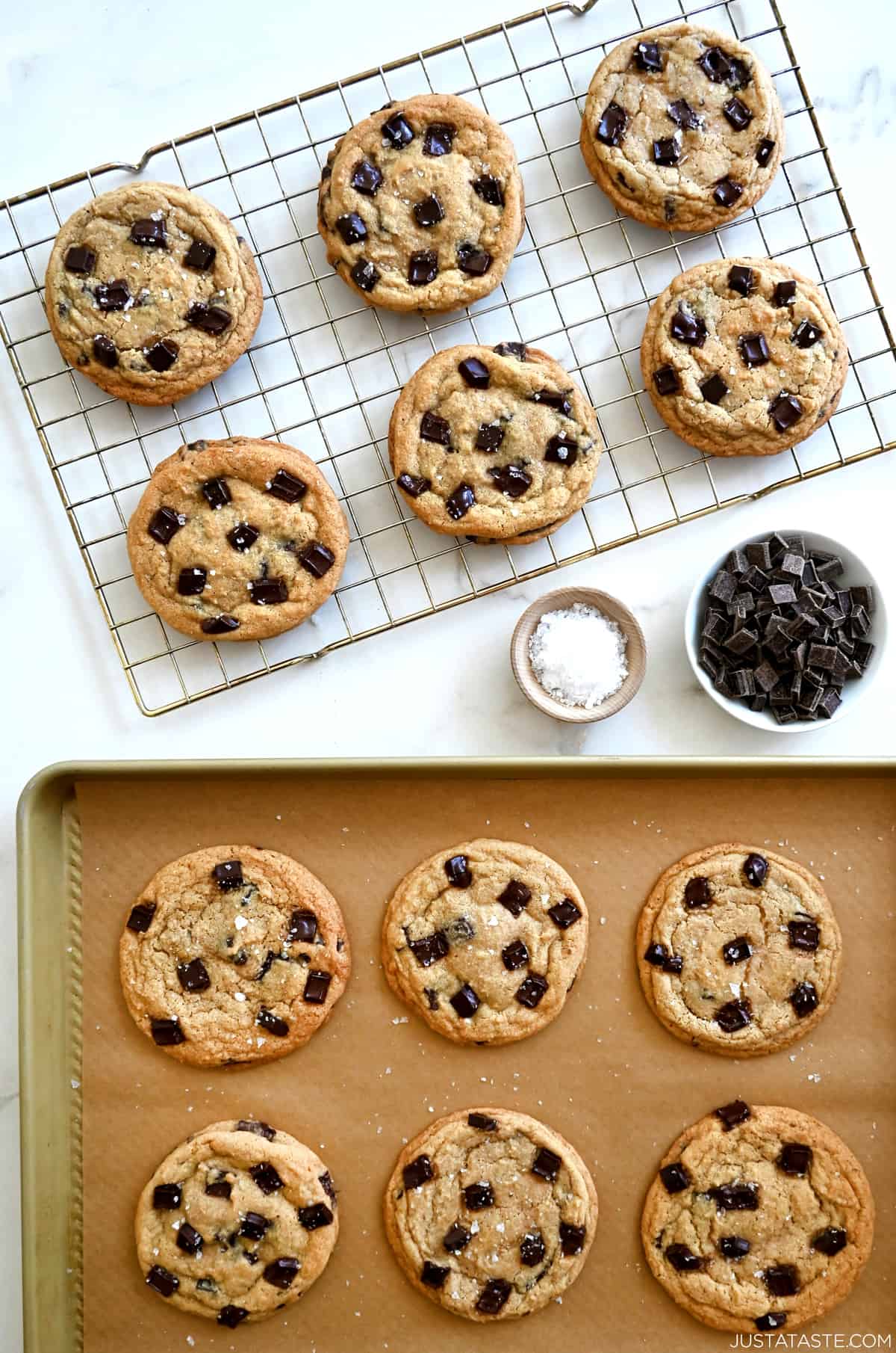 Chocolate chunk cookies on a wire baking rack next to freshly baked cookies topped with large-flake sea salt on a parchment paper-lined baking sheet.