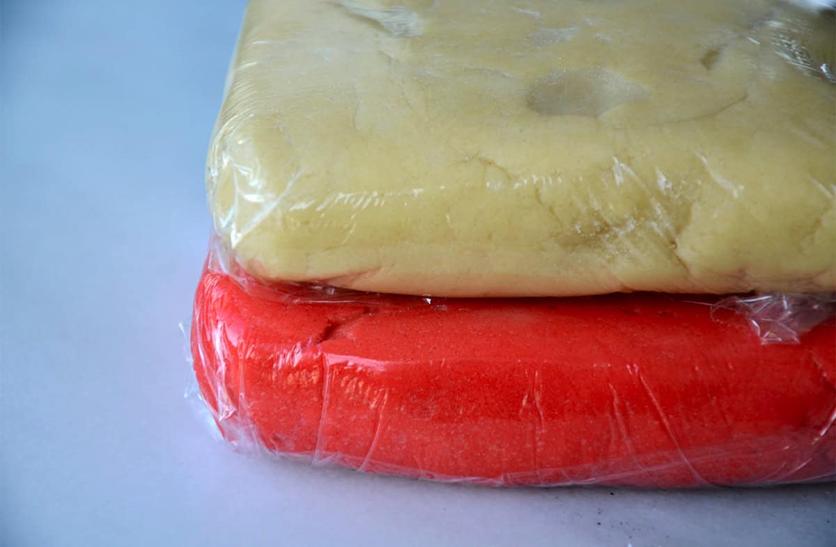 Pink sugar cookie dough and plain sugar cookie are wrapped in plastic wrap and stacked on a white countertop.