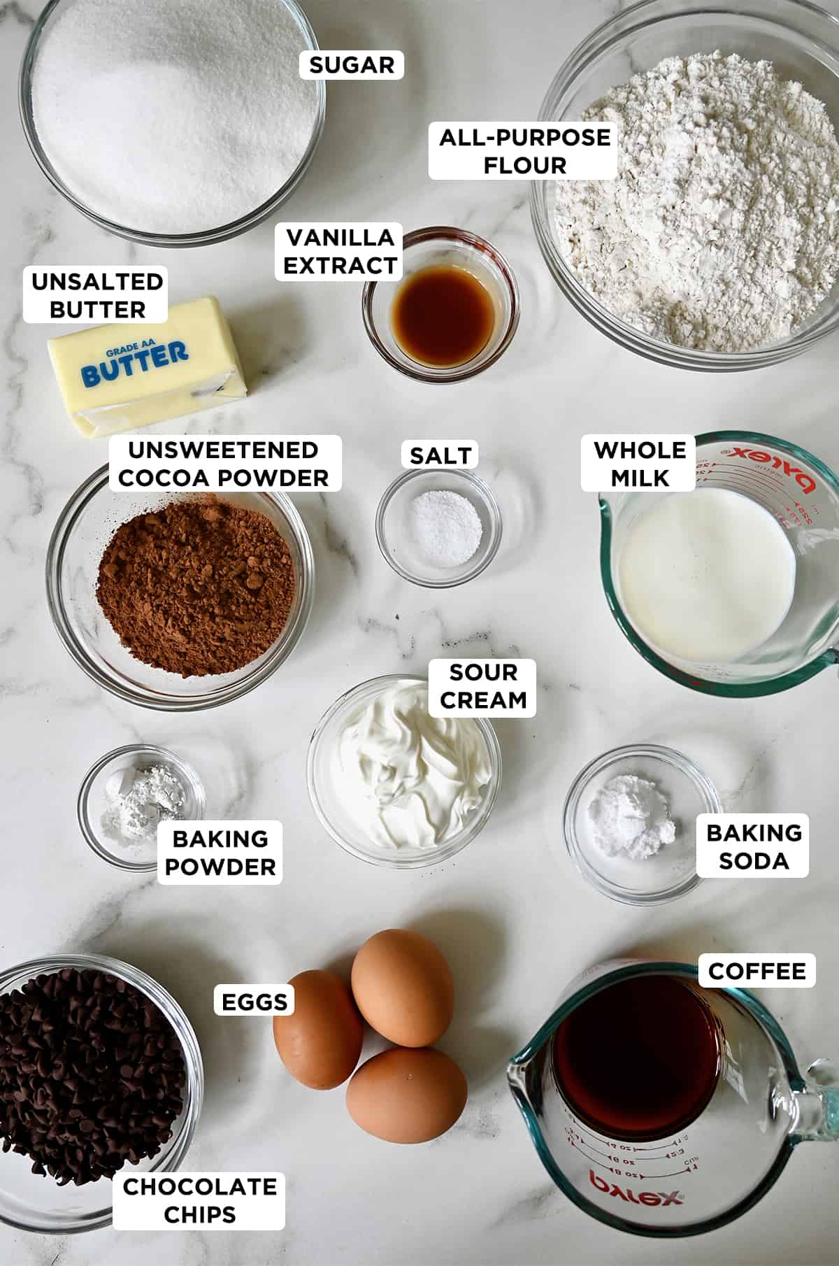 Chocolate cake ingredients in various sizes of glass bowls, including mini chocolate chips, baking powder, unsweetened cocoa powder, butter, sugar, vanilla extract, flour, salt, whole milk, sour cream, baking soda, coffee and three eggs.
