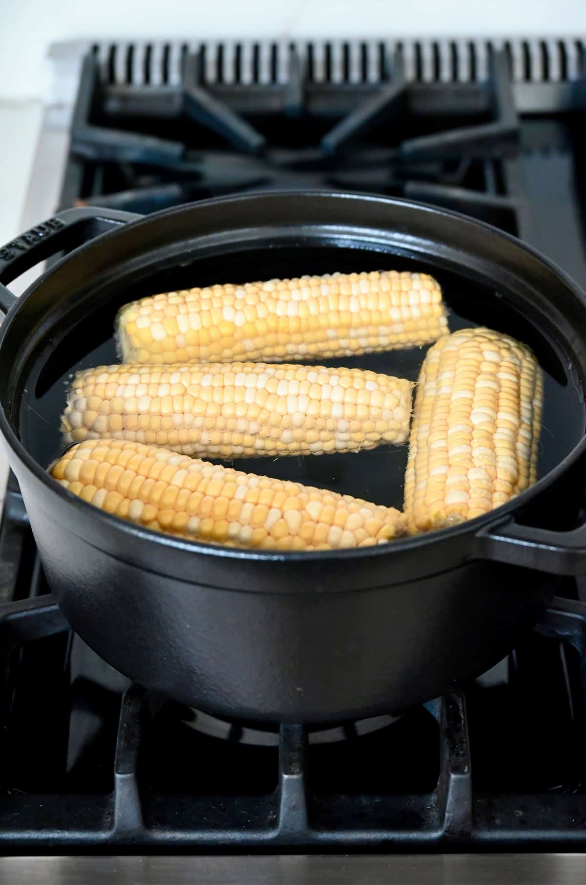 Four ears of shucked corn cooking in water in Dutch oven on a stovetop.
