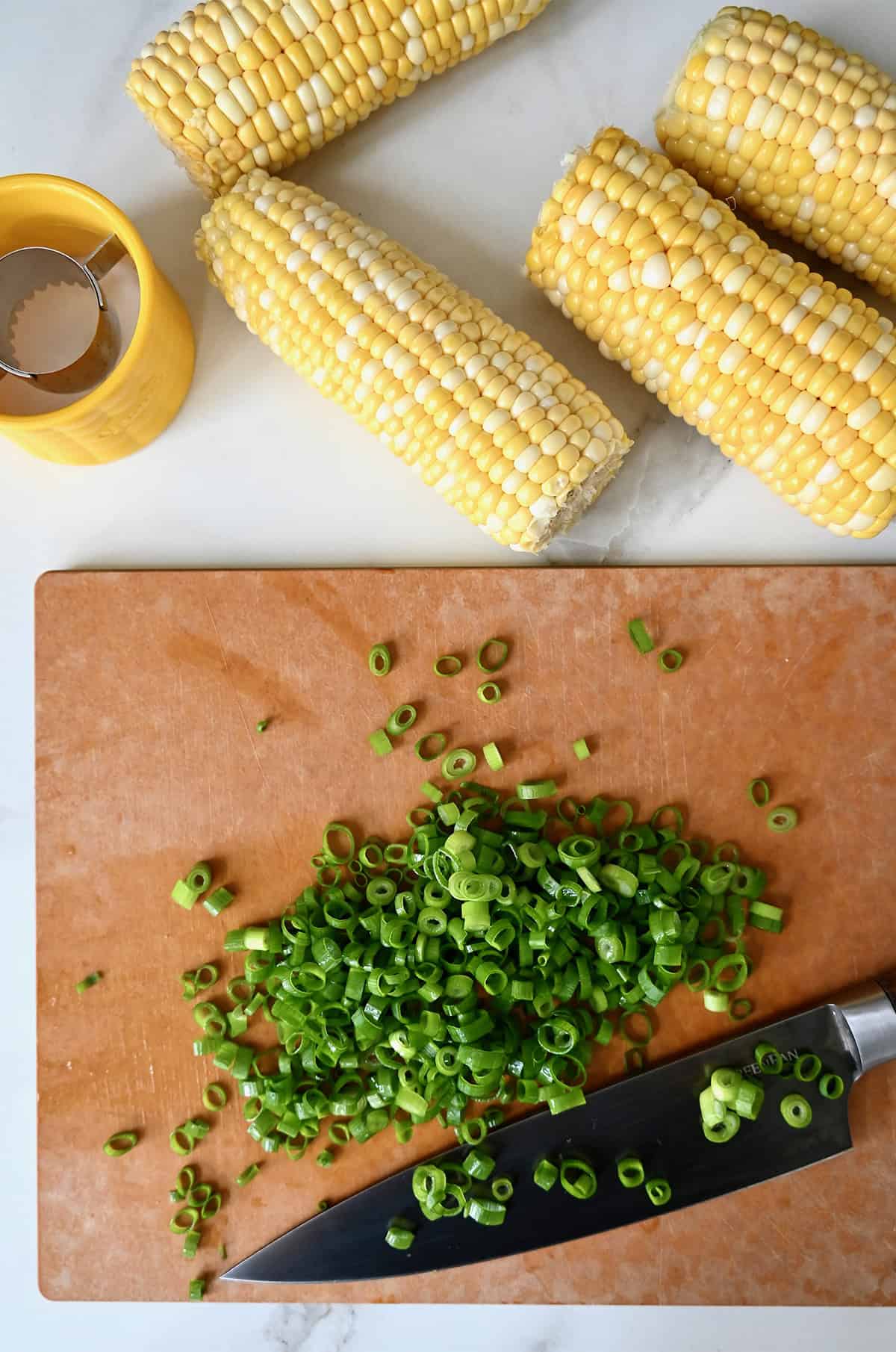 Thinly sliced scallions on a cutting board with a chef's knife. Four shucked ears of corn are beside the cutting board.