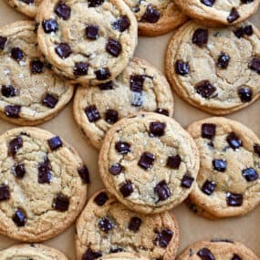 Dark chocolate chunk cookies with large-flake sea salt piled atop each other on brown parchment paper.