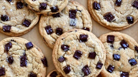 Dark chocolate chunk cookies with large-flake sea salt piled atop each other on brown parchment paper.