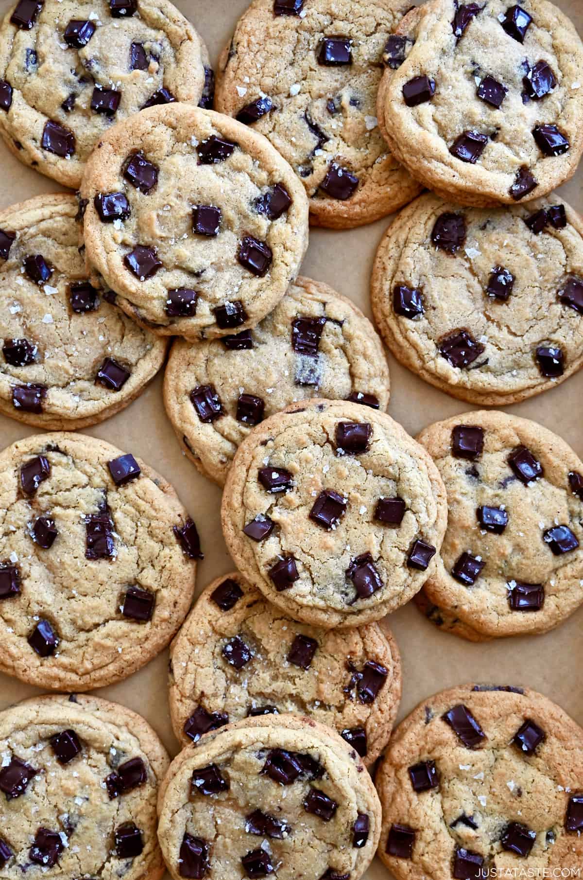 Dark chocolate chunk cookies with large-flake sea salt piled atop each other on brown parchment paper.