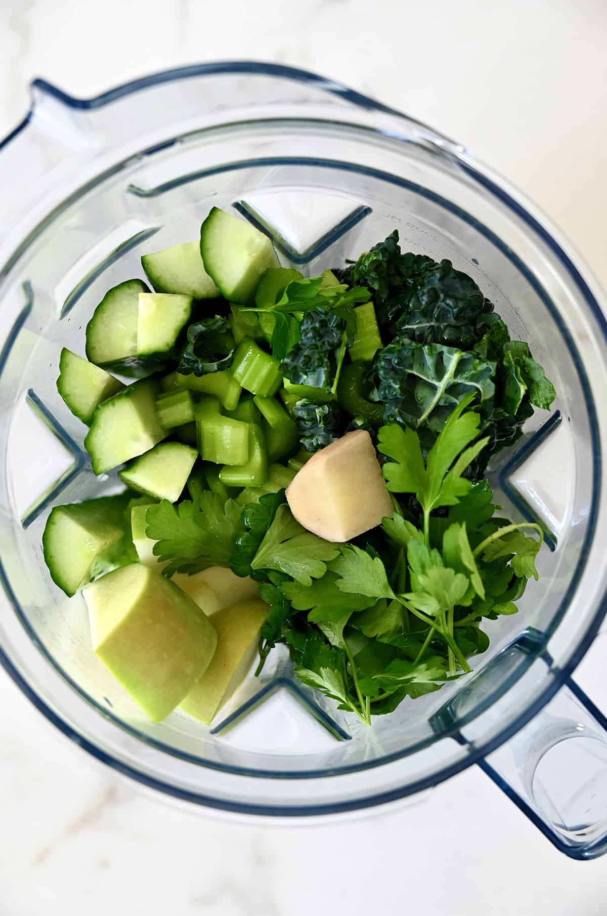 A blender jar is filled with ingredients for green juice, including cucumber, celery, ginger, apple and kale.