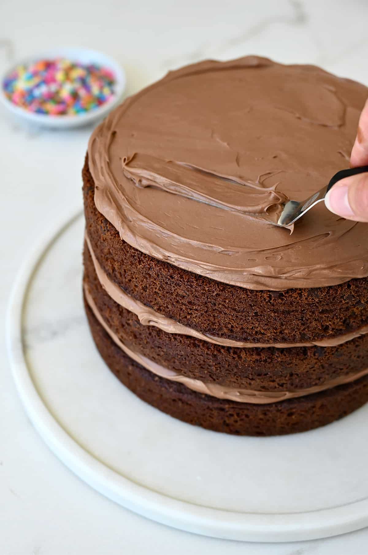 A hand holding an offset spatula spreads coffee buttercream to the top of a three-layer chocolate cake.