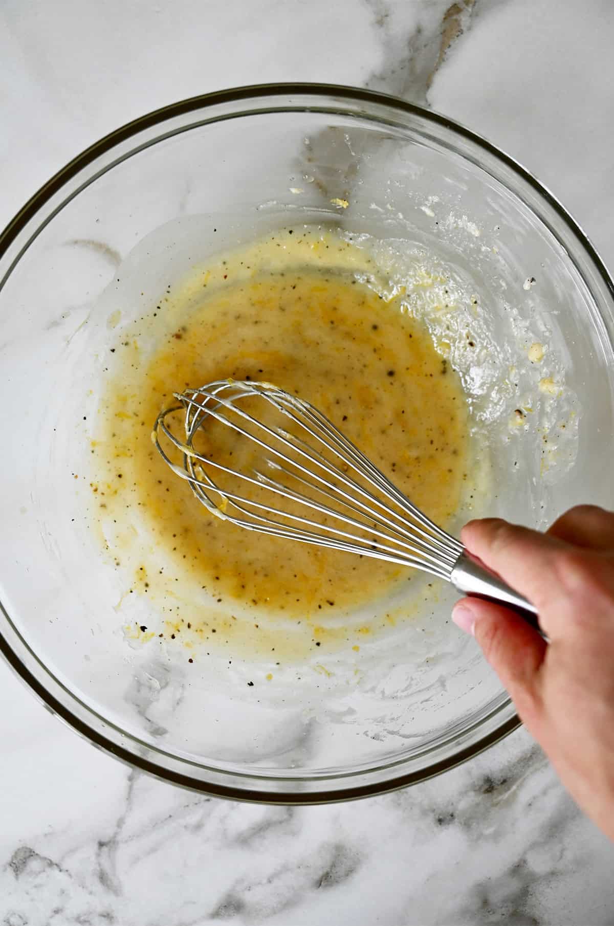 A person using a whisk to combine dressing in a large glass mixing bowl.