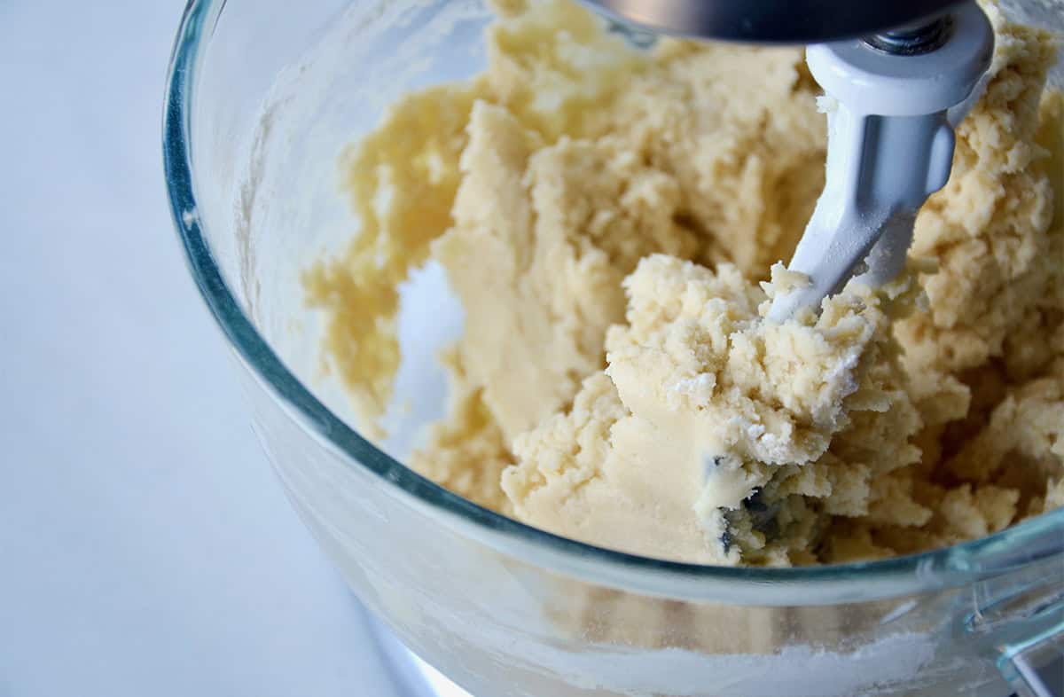 Sugar cookie dough is being mixed in the clear glass bowl of a stand mixer.