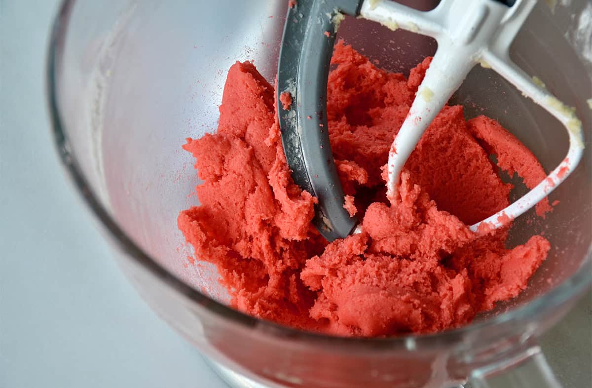 Pink sugar cookie dough is being mixed in a clear glass stand mixer bowl.