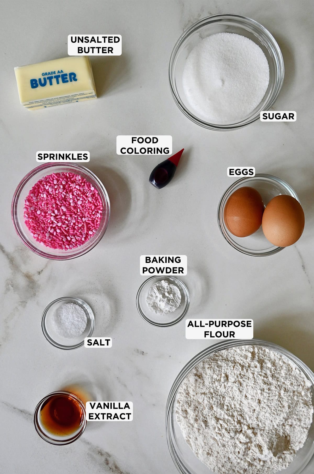 Ingredients for butter cookies are portioned into clear glass bowls, including flour, vanilla extract, salt, baking powder, eggs, sprinkles, food coloring, sugar and butter.