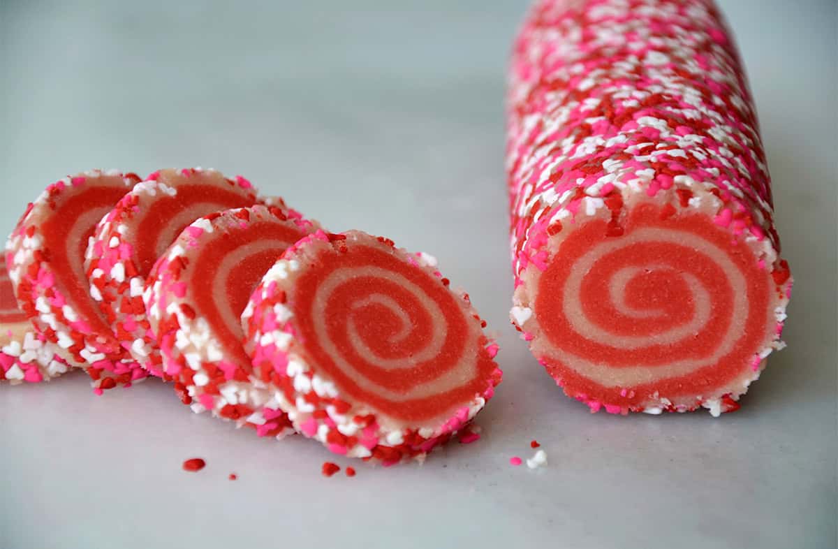 A log of pink and white pinwheel cookie dough, rolled in sprinkles, is cut into rounds.