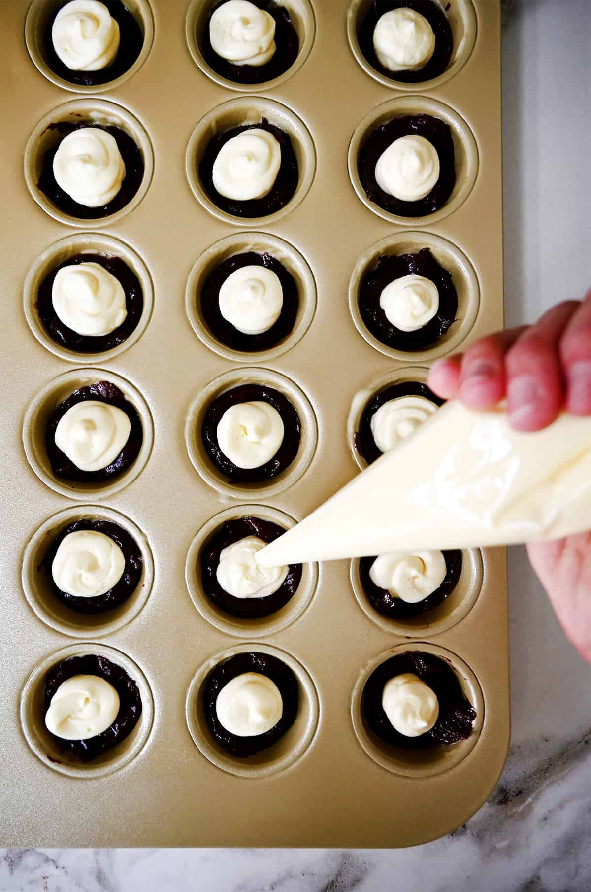 Cheesecake filling is piped onto brownie batter in a mini muffin pan.