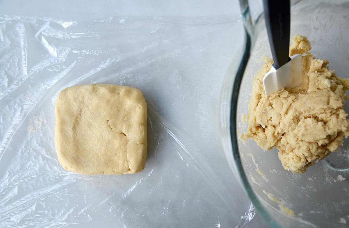 Sugar cookie dough is formed into a square on a sheet of plastic wrap, with a clear glass bowl of dough nearby.