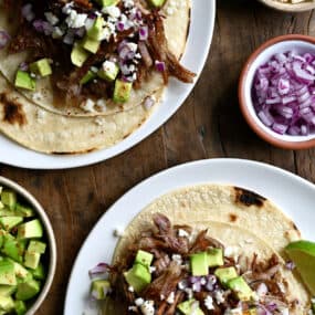 Two pulled pork tacos on separate plates. The tacos are topped with cubed avocado, crumbled cotija cheese and diced red onion. Small bowls with more toppings are beside the tacos.