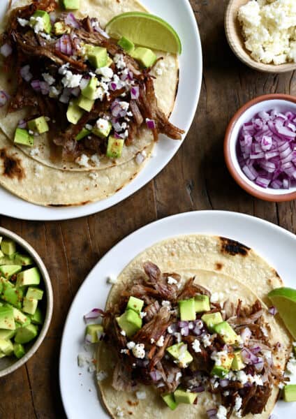 Two pulled pork tacos on separate plates. The tacos are topped with cubed avocado, crumbled cotija cheese and diced red onion. Small bowls with more toppings are beside the tacos.