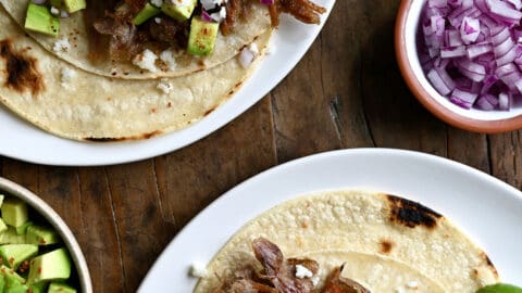 Two pulled pork tacos on separate plates. The tacos are topped with cubed avocado, crumbled cotija cheese and diced red onion. Small bowls with more toppings are beside the tacos.
