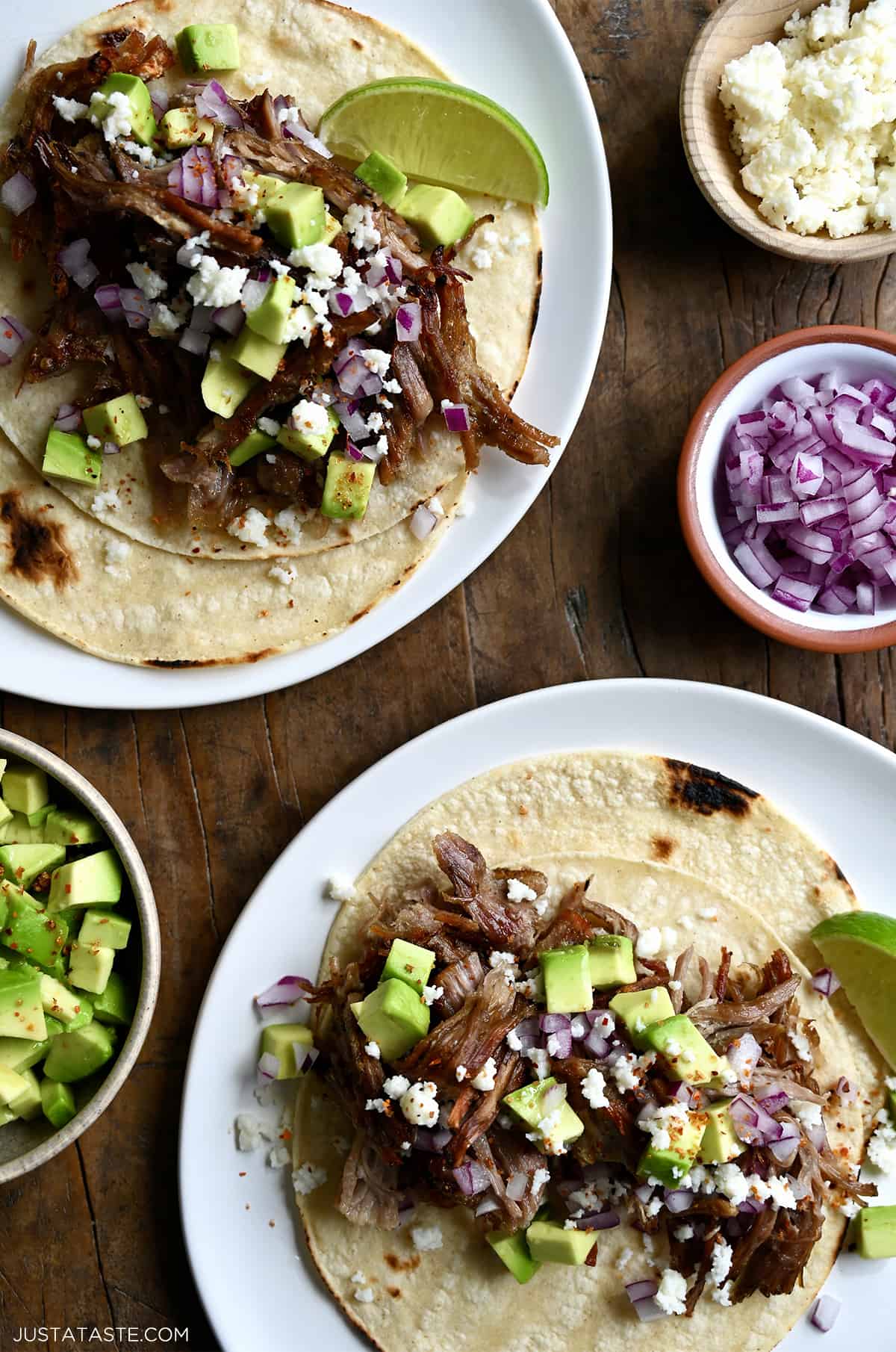 Two pulled pork tacos on separate plates. The tacos are topped with cubed avocado, crumbled cotija cheese and diced red onion. Small bowls with more toppings are beside the tacos.