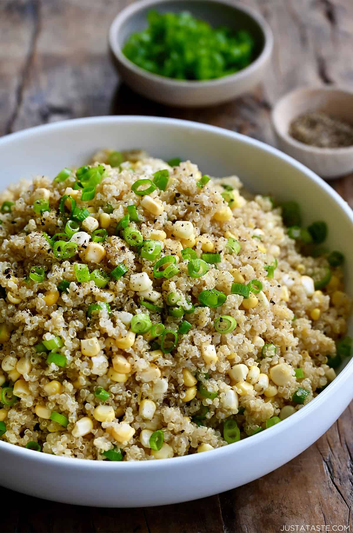 Quinoa with corn and scallions and topped with ground black pepper in a large white serving bowl. Small bowls of sliced scallions and ground black pepper are behind the serving bowl.