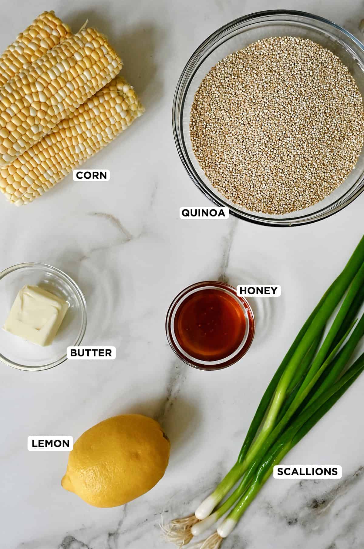 Four ears of shucked corn, three scallions, a lemon and small bowls of butter, honey and quinoa on a marble countertop.