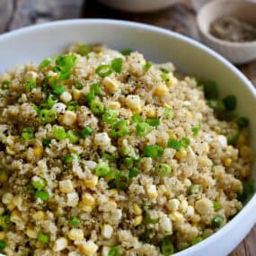 Quinoa with corn and scallions in a large white serving bowl. Small bowls of sliced scallions and ground black pepper are behind the serving bowl.