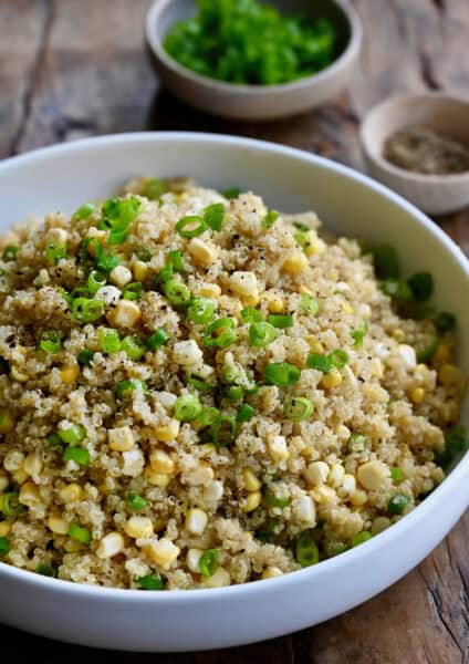 Quinoa with corn and scallions in a large white serving bowl. Small bowls of sliced scallions and ground black pepper are behind the serving bowl.