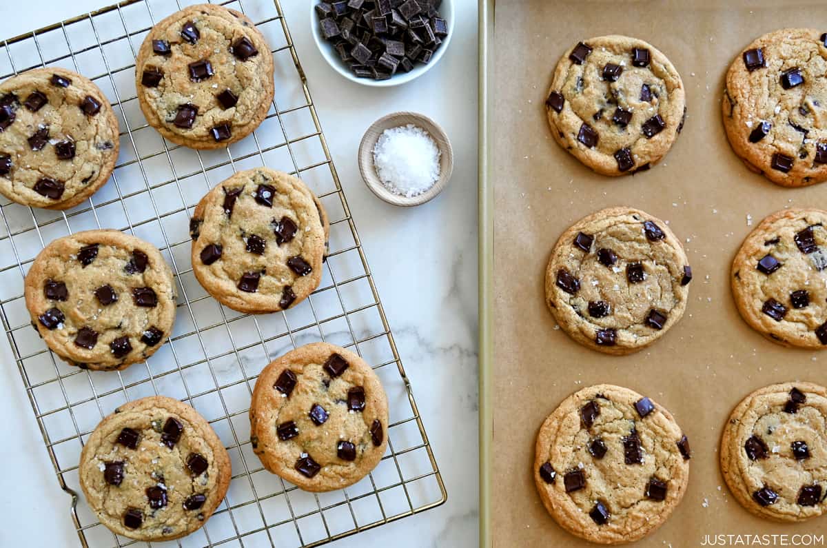 Chocolate chunk cookies cooling on a wire rack next to freshly baked dark chocolate chunk cookies on a parchment paper-lined baking sheet. A small bowl filled with large-flake sea salt and a small bowl containing chocolate chunks are nearby.