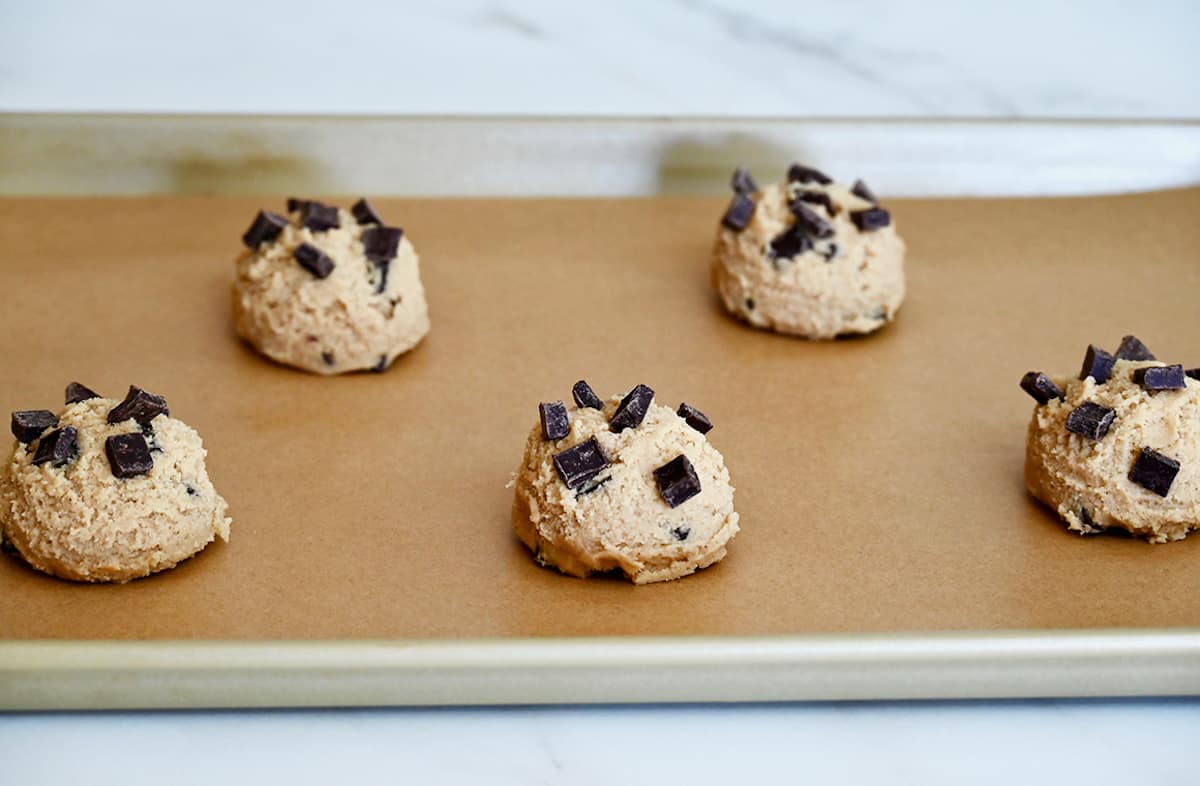 Balls of cookie dough topped with chocolate chunks on a parchment paper-lined baking sheet.