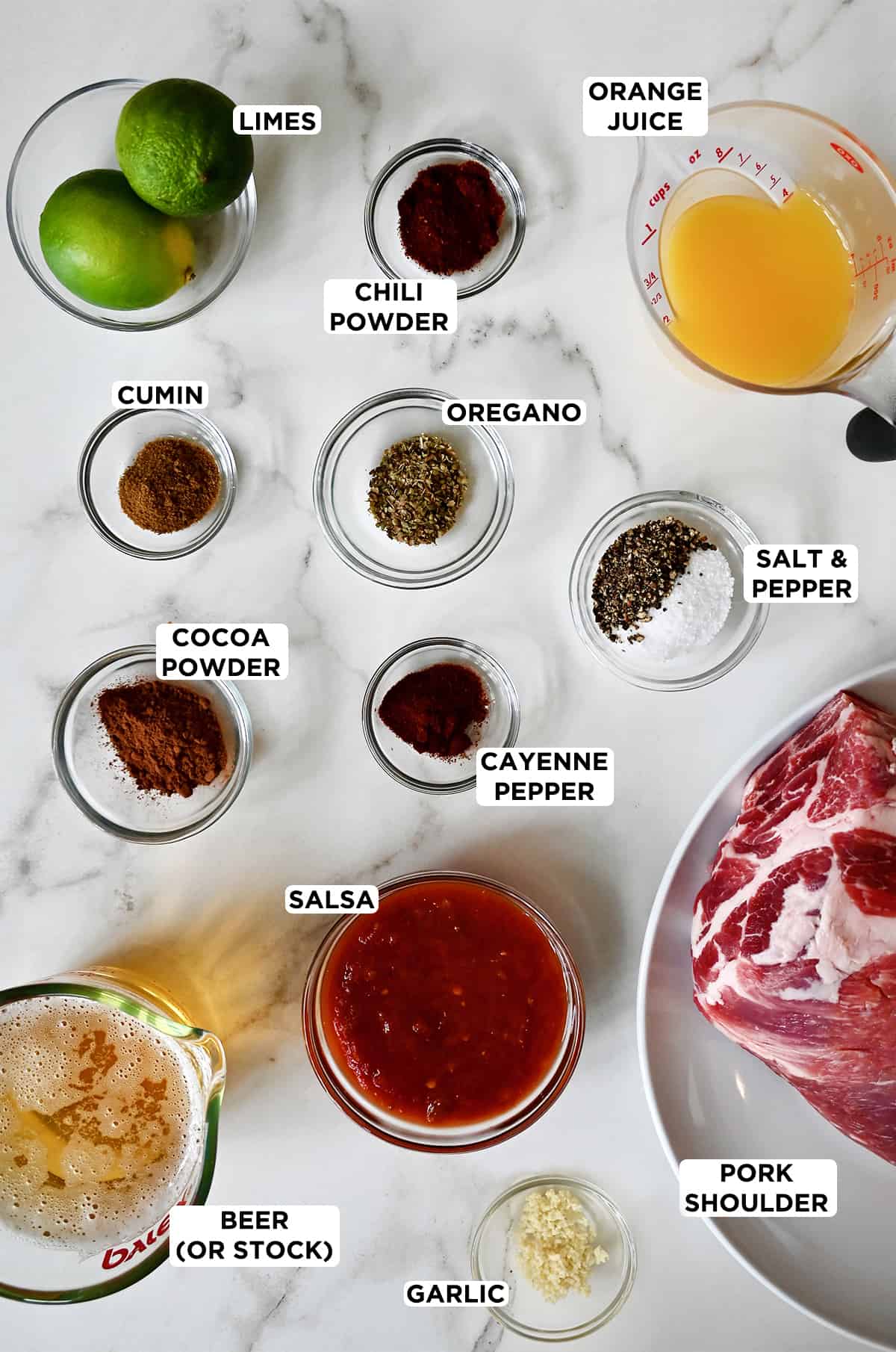 Small bowls of limes, chili powder, oregano, cumin, salt and pepper, cocoa powder, cayenne pepper, salsa and minced garlic on a marble countertop. Measuring cups containing beer and orange juice and plate with a pork shoulder on it are also on the countertop.