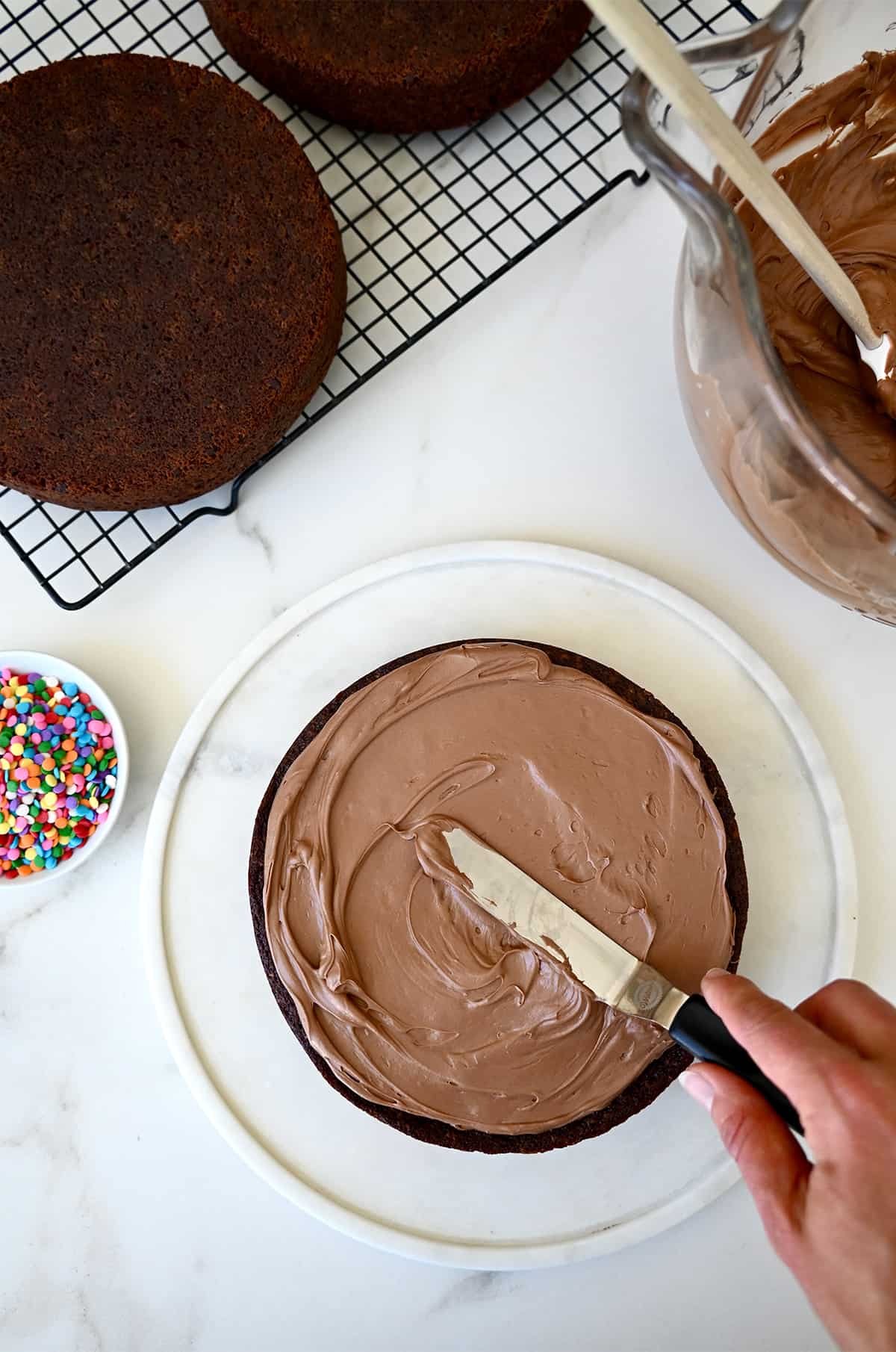 A hand holding an offset spatula spreads coffee buttercream frosting atop a chocolate cake.
