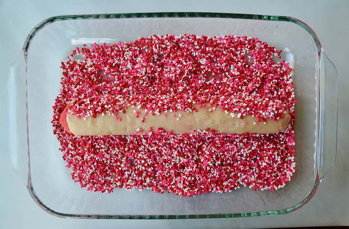 A log of slice and bake sugar cookies is rolled in pink, white and red sprinkles in a clear glass dish.