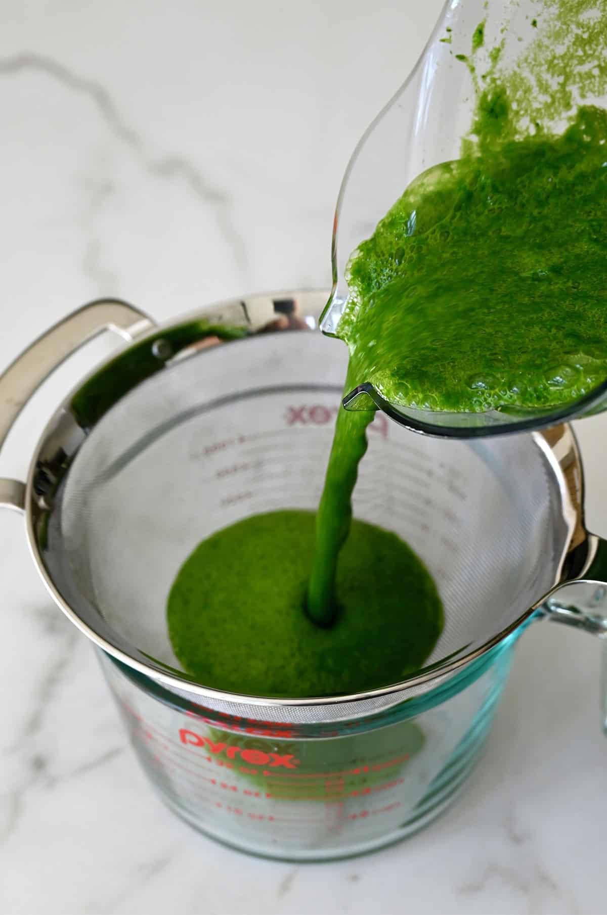 Green juice is poured through a fine mesh strainer into a glass measuring cup.